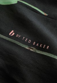 Black fabric featuring a green stem and leaf graphic, with the text "b by TED BAKER" in light pink below. Smooth texture and subtle sheen.