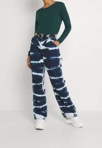Grünes Langarmoberteil mit einem strukturierten Muster, kombiniert mit blauen Batik-Hosen mit weitem Bein, die weiße Spiralen und einen schwarzen Gürtel aufweisen. Weiße Sneaker.