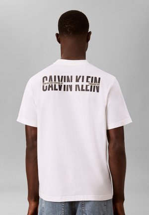 Άνδρας που φοράει λευκό t-shirt Calvin Klein με λογότυπο και κείμενο στο πάνω μέρος της πλάτης, σε συνδυασμό με μπλε τζιν, στέκεται μπροστά σε γκρι φόντο.