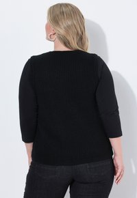 Svart stickad väst med ribbat mönster, bärs över en svart topp, tillsammans med mörka jeans. Anmärkningsvärt inslag inkluderar stora pärlliknande örhängen.