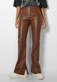 Pantaloni a zampa d'elefante in finta pelle marrone con vita alta, caratterizzati da spacco laterale all'orlo e una texture liscia e lucida. Combinati con sneakers bianche.