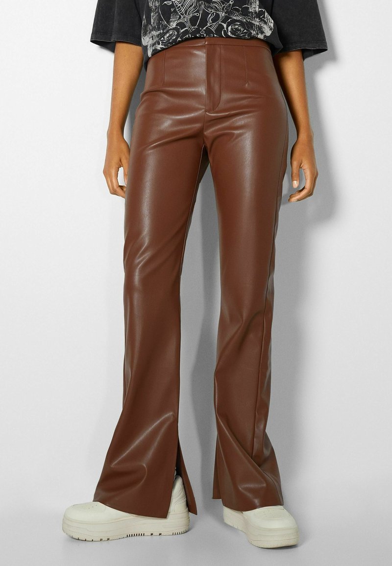 Pantaloni a zampa d'elefante in finta pelle marrone con vita alta, caratterizzati da spacco laterale all'orlo e una texture liscia e lucida. Combinati con sneakers bianche.