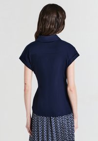 Donna con lunghi capelli castani che indossa una blusa azzurra a maniche corte e una gonna a righe blu e bianca, in piedi con le spalle rivolte verso la fotocamera.