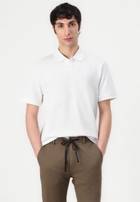 Hvit poloshirt med korte ermer, med en klassisk krage og en subtil logo. Kombinert med olivengrønne joggebukser med snøring, som har en avslappet passform.