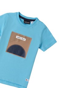 Camiseta de algodón turquesa con un parche redondo azul marino. Gráfico rectangular marrón que presenta el texto "NO ESPERES" en blanco, con texto repetido.