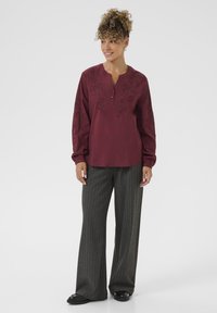Bordeaux blouse met geborduurd bloemenontwerp, knoophalslijn, lange mouwen; gecombineerd met grijze broek met fijne strepen en zwarte schoenen.