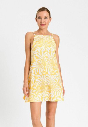 Donna che indossa un mini abito giallo e bianco senza maniche con sottili bretelle, in piedi davanti a uno sfondo chiaro e uniforme.