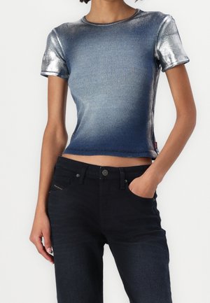 Frau trägt ein metallisch blaues, eng anliegendes Kurzarm-Oberteil und schwarze Hosen, steht mit einer Hand in der Tasche vor neutralem Hintergrund.
