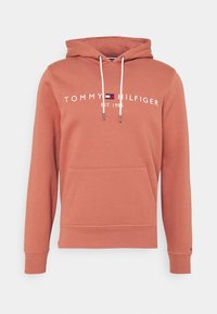 Rust-colored hoodie with front pocket, white drawstrings, and "Tommy Hilfiger Est. 1985" logo in white and navy red on the chest.