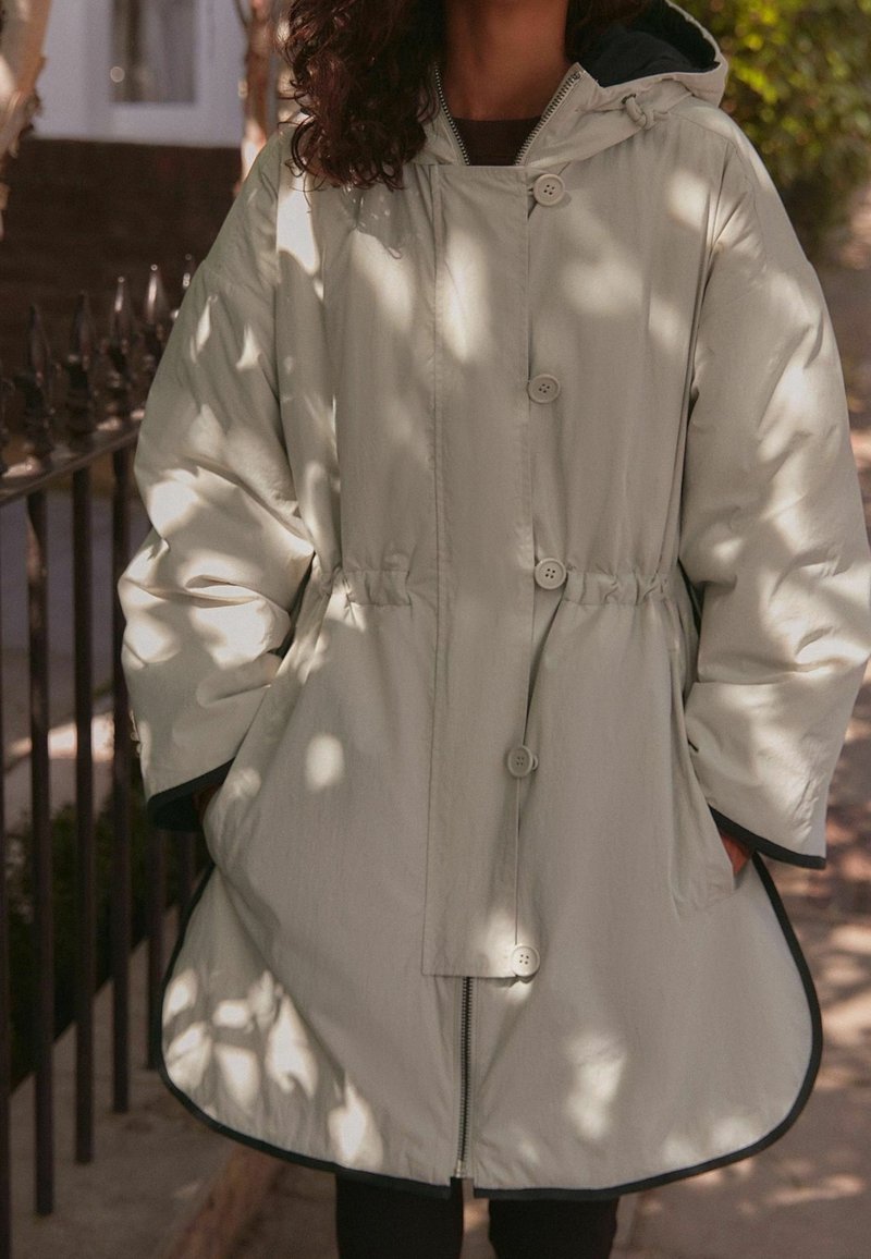 Femme portant un manteau à capuche beige clair avec boutons et fermeture éclair, mains dans les poches, debout à l'extérieur près d'une clôture métallique noire.