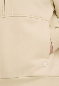 Beige sweatshirt with a front pocket, ribbed cuffs, and a small logo detail. Made from soft, textured fabric, featuring a half-zip design.