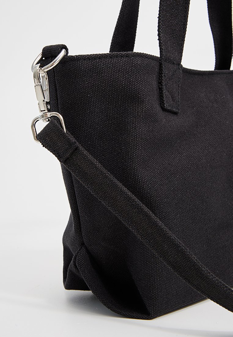 Sac fourre-tout en toile noire avec une bandoulière amovible accrochée à un anneau en métal dans le coin supérieur, sur fond blanc.