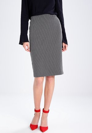 Jupe crayon à pois noir et blanc avec une coupe ajustée. Portée avec un haut noir à manches longues et des chaussures à talons rouges.
