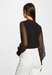 Top nero con lunghe maniche trasparenti a fantasia, caratterizzato da un collo rotondo e un corpino aderente, abbinato a pantaloni chiari a vita alta.