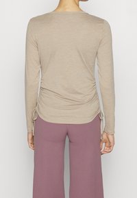 Long-sleeve beige top with gathered side detail and adjustable ties, paired with loose-fitting mauve pants. Smooth texture, simple design.