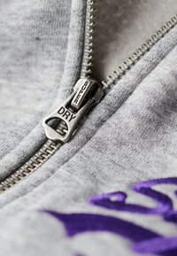 Tessuto grezzo grigio con zip metallica; il cursore della zip è contrassegnato con "SUPER DRY". Dettaglio ricamato viola accanto alla zip.