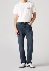 Vit kortärmad t-shirt med färggloggigt textlogo, kombinerad med mörkblå raka denimjeans och vita sneakers med svarta detaljer.