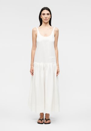 Γυναίκα που φοράει λευκό μάξι φόρεμα χωρίς μανίκια με φραμπαλά φούστα και μαύρα σανδάλια, στέκεται μπροστά σε ένα μονόχρωμο ανοιχτό φόντο.