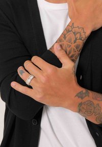 Bague en argent avec un motif gravé, portée sur une main tatouée. Cardigan noir et T-shirt blanc visibles, mettant en valeur le teint de peau.