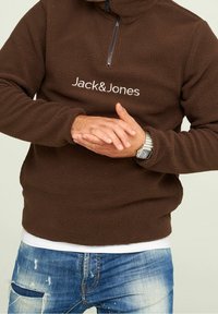 Sudadera de felpa marrón con cuello de media cremallera, con el logotipo blanco "Jack & Jones" en el pecho. Combinada con unos vaqueros azules desgastados.