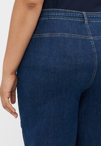 Jeans de denim azul escuro com uma textura suave, apresentando uma cintura média e detalhes sutis de costura ao longo das costuras.
