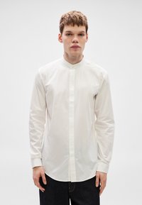 Λευκό πουκάμισο με μακριά μανίκια και γιακά μπάντας, με κλείσιμο με κουμπιά στο μπροστινό μέρος και λεία υφή. Φορεμένο με σκούρο τζιν.