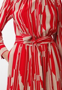 Gros plan sur une robe rayée rouge et beige avec un motif abstrait, ceinturée à la taille avec une ceinture en tissu assortie, main dans la poche.