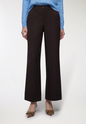 Donna che indossa pantaloni neri a gamba larga e cropped, un maglione azzurro chiaro e scarpe a punta marroni con dettagli in fiocco e borchie.