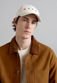 Căciulă albă de baseball cu text roșu "MARNI," jachetă maro din catifea cu o guler neted și cămașă bej dedesubt. Apăreație curată, minimalistă.