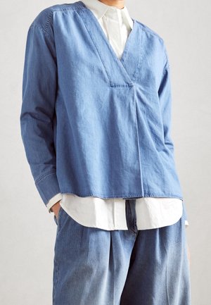 Person iført lagdelt, løs blå denimtop over hvid skjorte med krave, kombineret med matchende bredbenede denimbukser, stående foran en ensfarvet baggrund.