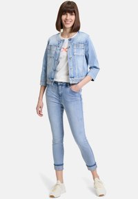 Ljust blå denimjacka med två framfickor och rund halsringning, tillsammans med ljusblå åtsittande jeans. Modellen har på sig en vit t-shirt och sneakers.