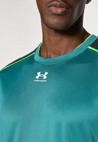 Türkizkék sportpóló kerek nyakkivágással, zöld díszítésekkel és fekete vállrésszel. Az Under Armour logó a mellrészen. Simább, szintetikus anyagból készült.