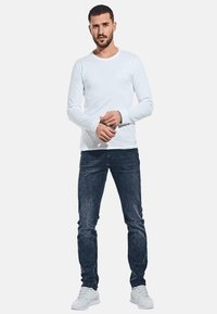 Weißes, langärmeliges Hemd kombiniert mit dunkelblauen Jeans und weißen Turnschuhen. Das Hemd hat einen runden Ausschnitt und enganliegende Ärmel.