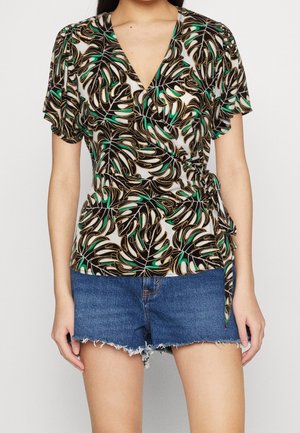 Femme portant un chemisier cache-cœur à imprimé de feuilles tropicales avec manches ballons et un short en jean bleu coupé sur un fond uni.