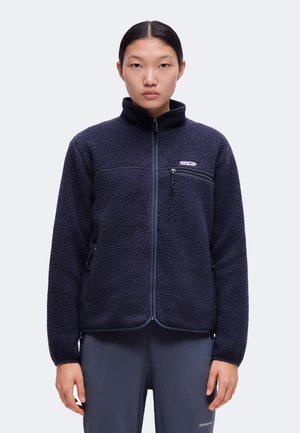 Νεαρό άτομο φοράει σκούρο μπλε fleece μπουφάν με τσέπη στο στήθος με φερμουάρ και ταιριαστό παντελόνι, σε απλό φωτεινό φόντο.
