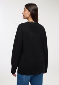 Kobieta z długimi ciemnymi włosami, ubrana w czarny sweter o fakturze i niebieskie dżinsy, stoi bokiem na tle białego tła.