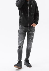Black bomber jacket with zippered pockets, paired with distressed gray skinny jeans featuring rips and text detail. Black high-top sneakers.