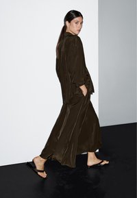 Donkergroene zijden maxi-jurk met brede mouwen en zijzakken, gecombineerd met platte bruine sandalen, tegen een minimalistische achtergrond.