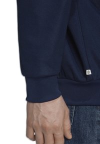 Mörk marinblå sweatshirt med ribbstickad mudd, en subtil logotypetikett i fållen och ett strukturerat tyg, delvis synligt på en hand.