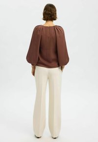 Blusa marrone a maniche lunghe con maniche a sbuffo, dettaglio a chiave sulla schiena, abbinata a pantaloni a gamba larga di colore crema. Texture complessivamente liscia.
