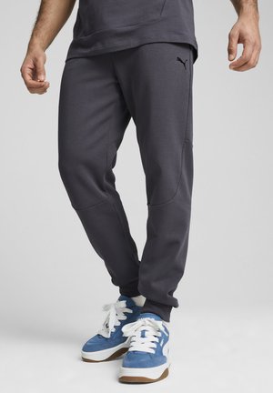 Pantalones deportivos de color gris oscuro con piernas ajustadas, bolsillos laterales, logo en la cadera, combinados con zapatillas azules que tienen cordones blancos y una suela blanca.