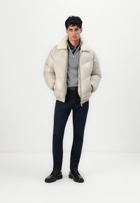 Beige pufferjack met een fluffy kraag, gedragen over een grijze trui en een marineblauwe shirt, gecombineerd met marineblauwe broek en zwarte veterschoenen.