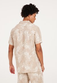 Camisa de manga corta en beige claro con un patrón de hojas blancas, que presenta un cuello clásico y un corte relajado, hecha de una tela suave y texturizada.