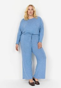 Ljusblå, avslappnad jumpsuit i mjukt tyg med långa ärmar och dragsko i midjan. Matchad med svarta, vävda skor med öppen tå.