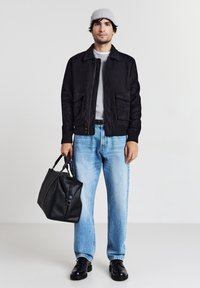 Giacca di suede nera con tasche frontali, maglione grigio, jeans blu sbiaditi, borsa di pelle nera e scarpe nere. Il modello indossa un berretto grigio.