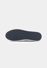 Suela de zapato de goma oscura con una superficie texturizada negra, borde blanco y una etiqueta de logo rectangular que dice "TOMMY HILFIGER" en el centro.