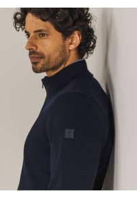 Dunkelblau gestrickter Pullover mit hohem Kragen, aus strukturiertem Stoff, in schmaler Passform und mit einem kleinen Markenpatch an der linken Ärmeln.