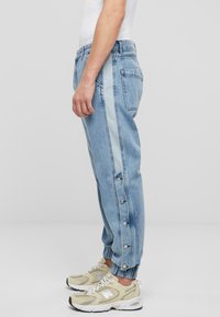 Pantaloni din denim albastru deschis, cu o croială lejeră, având nasturi cu prindere tip snap de culoare argintie pe margini și un panou lateral într-o nuanță ușor mai deschisă de albastru.