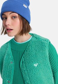 Turkusowa kamizelka z polaru o teksturowanej powierzchni, nałożona na morski sweter z dzianiny. Stylizację dopełnia niebieska prążkowana czapka.