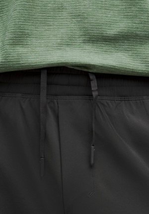 Maglietta atletica verde con finitura testurizzata, indossata su pantaloni corti neri con vita regolabile e tessuto liscio.